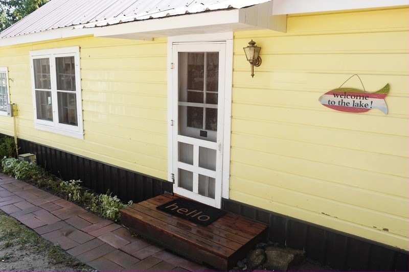 The pale yellow cabin, its white screen door, a hello mat and a painted fish sign reading welcome to the lake